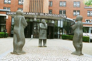 Das Bild zeigt drei antropomorphe Skulpturen vor dem Bildungszentrum der Handwerkskammer. Die Figuren stehen einander gegenüber und wirken nachdenklich.