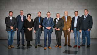 Das Bild zeigt eine Gruppe von neun Personen in formeller Kleidung, die auf einer modernen Oberfläche vor einer grauen Wand stehen.